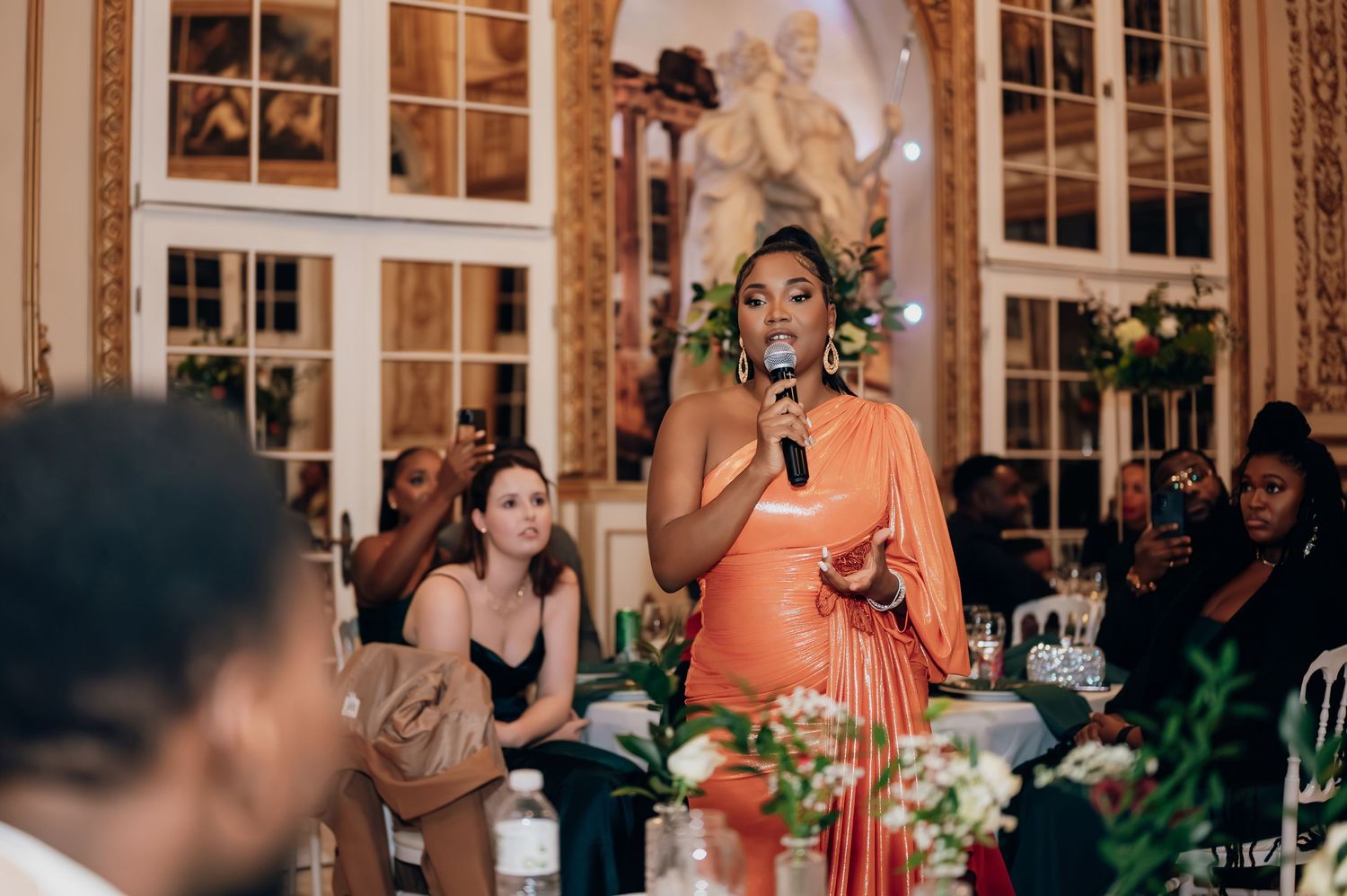 Woman in elegant orange one-shoulder gown delivering a heartfelt speech at a wedding reception.