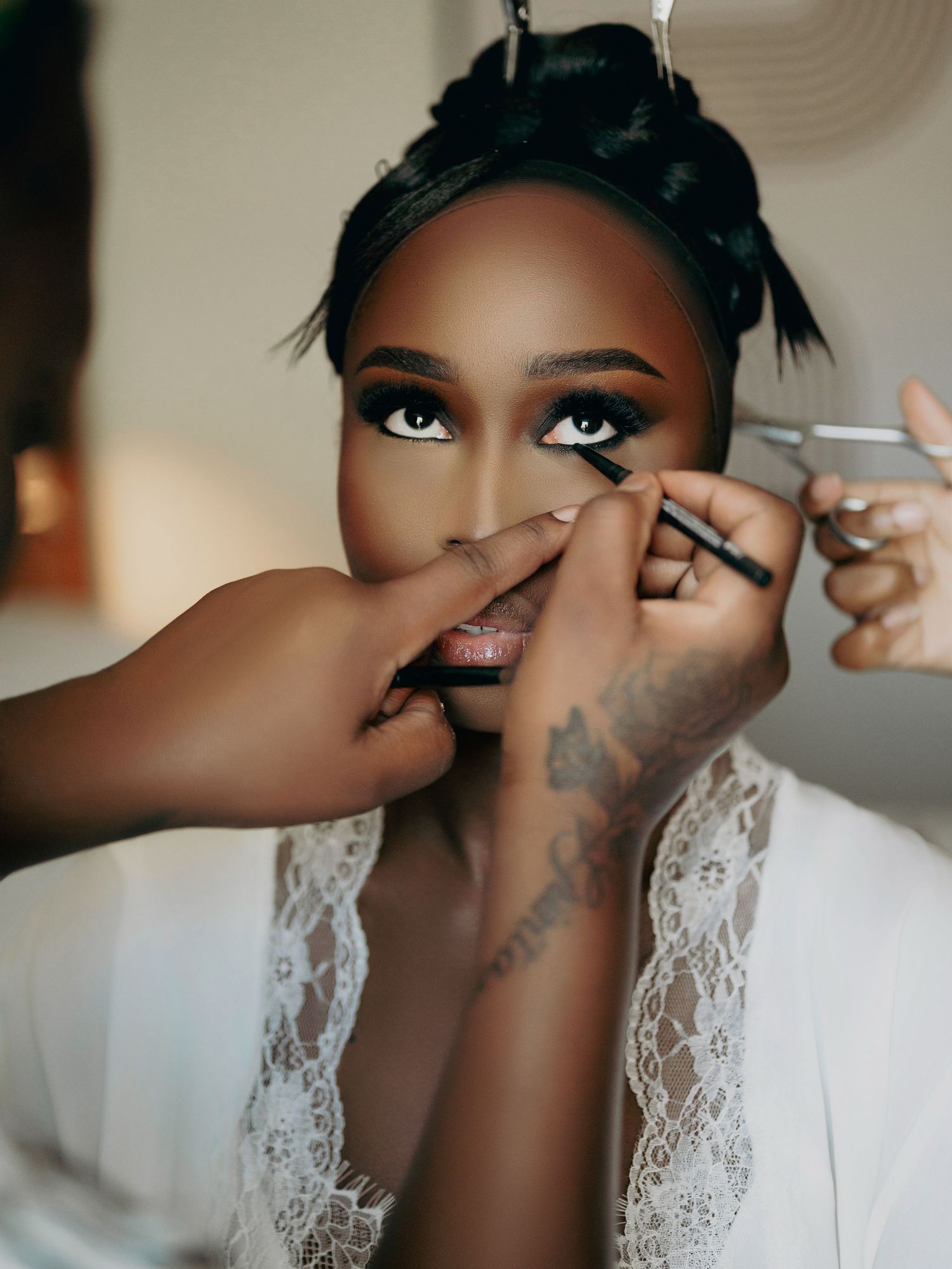Makeup artist applying eyeliner to a bride in a white lace robe.