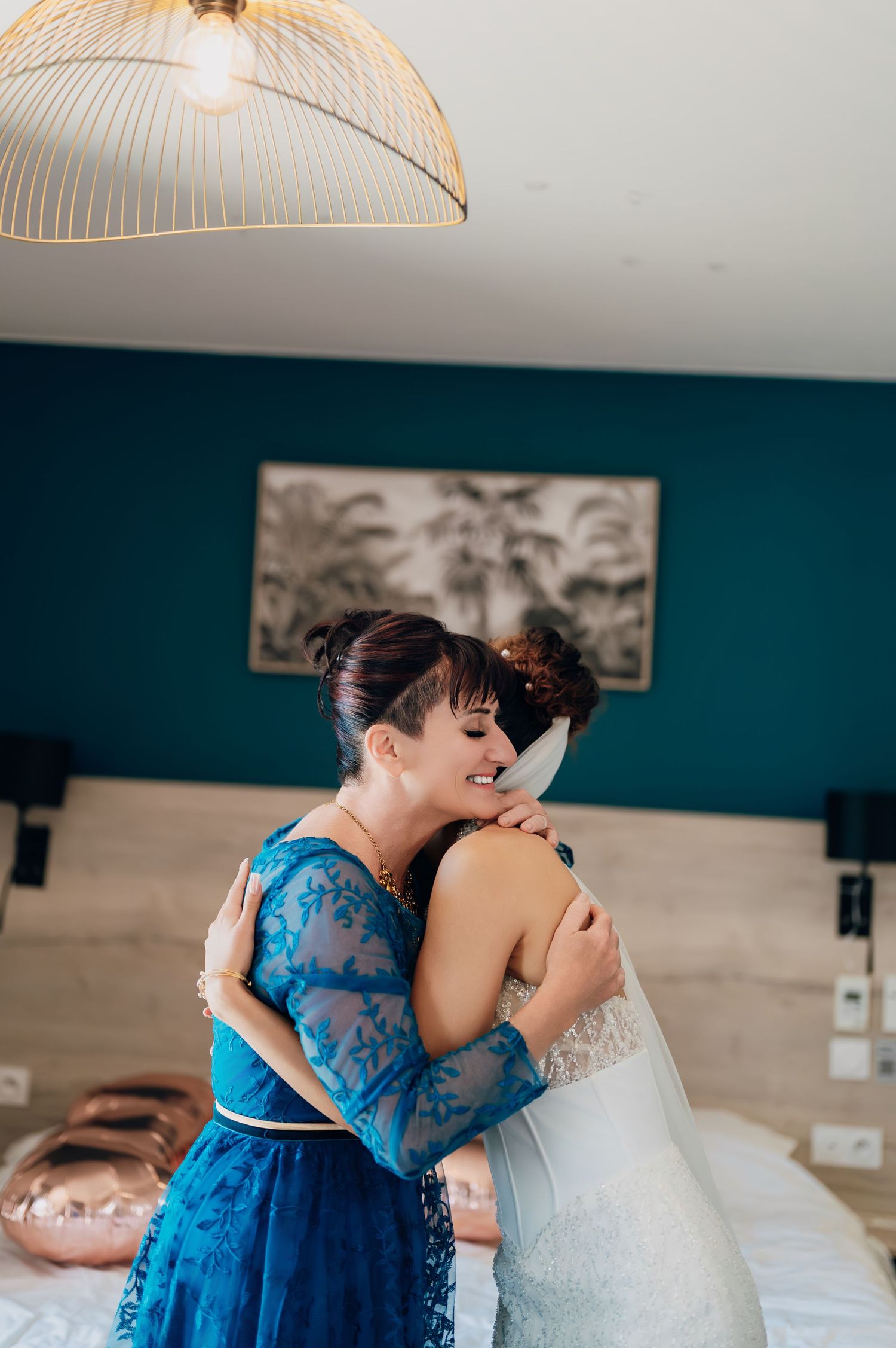 Emotional mother and bride share a warm embrace in a teal-walled hotel room on the wedding morning.