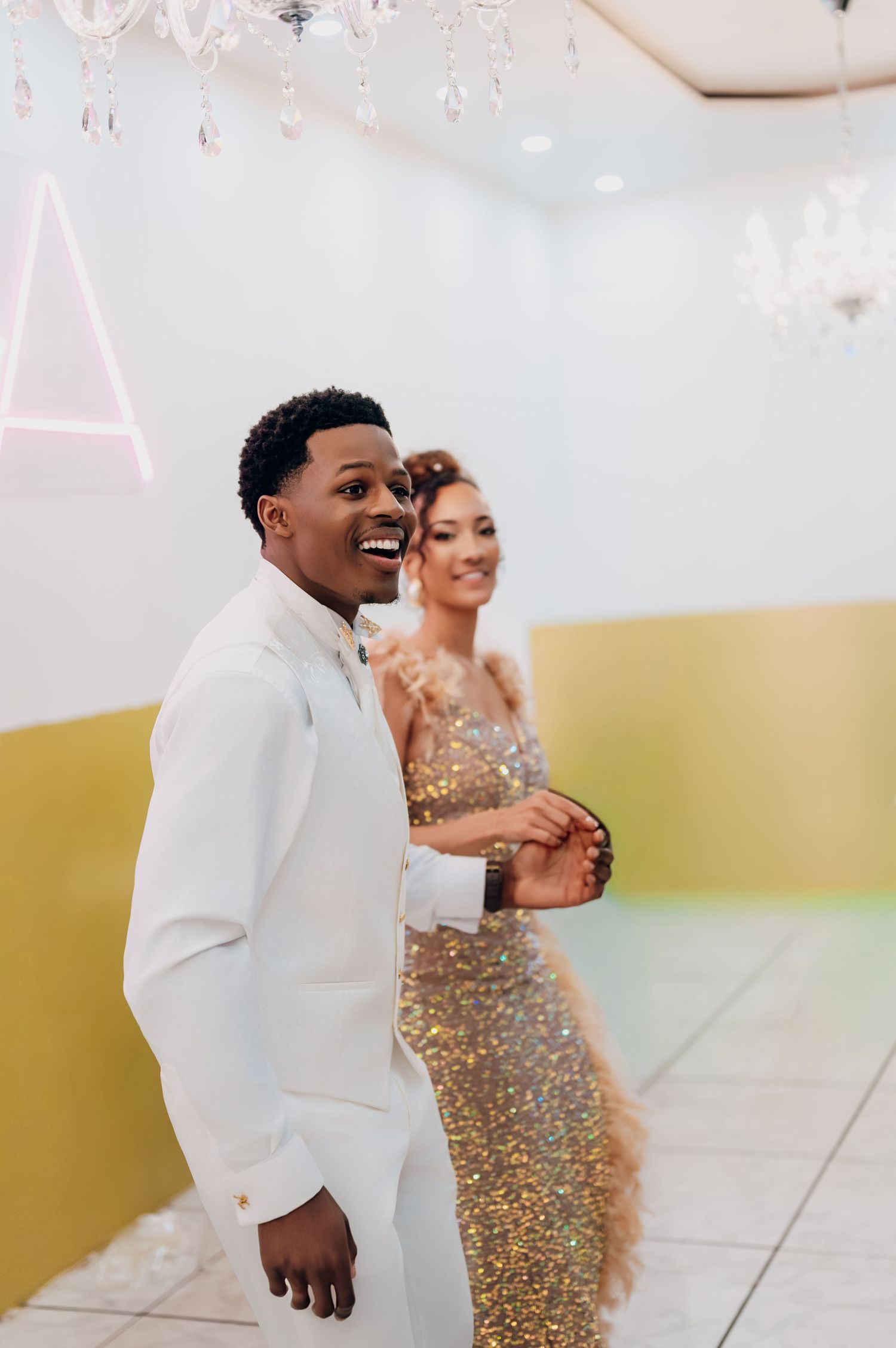 Smiling groom in white suit leads bride in gold sequin dress through wedding reception venue.