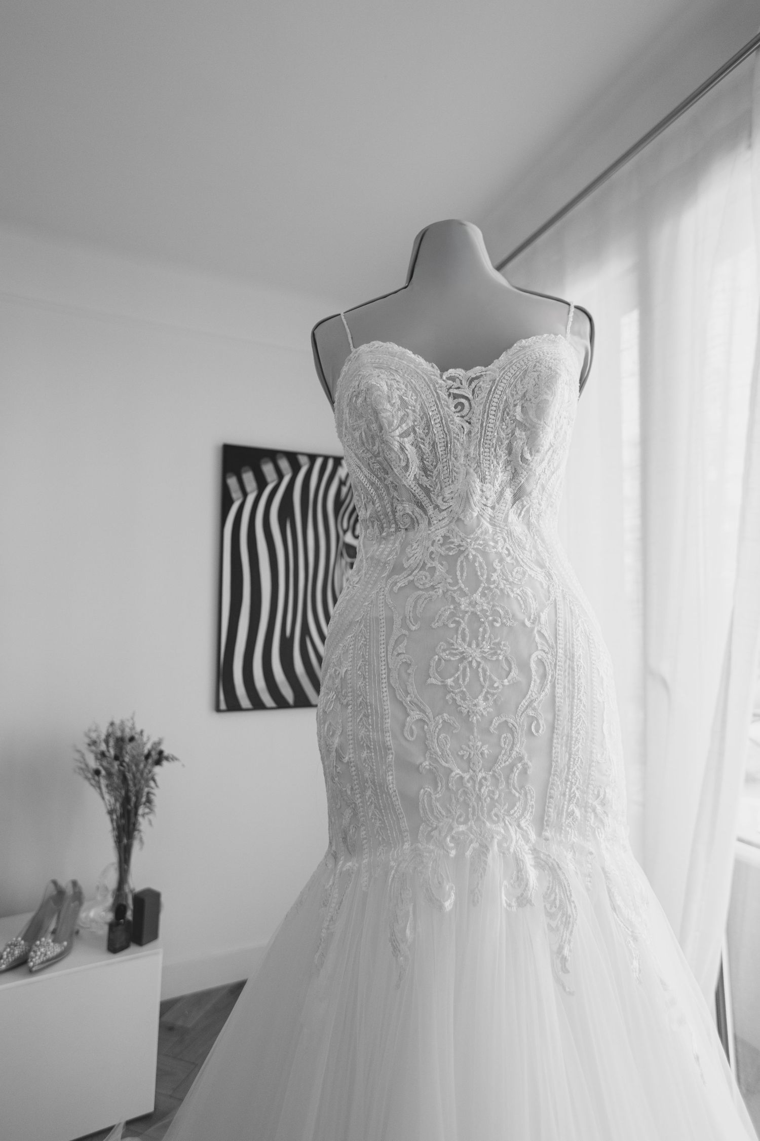 Black and white photo of elegant lace mermaid wedding dress displayed on mannequin near window.