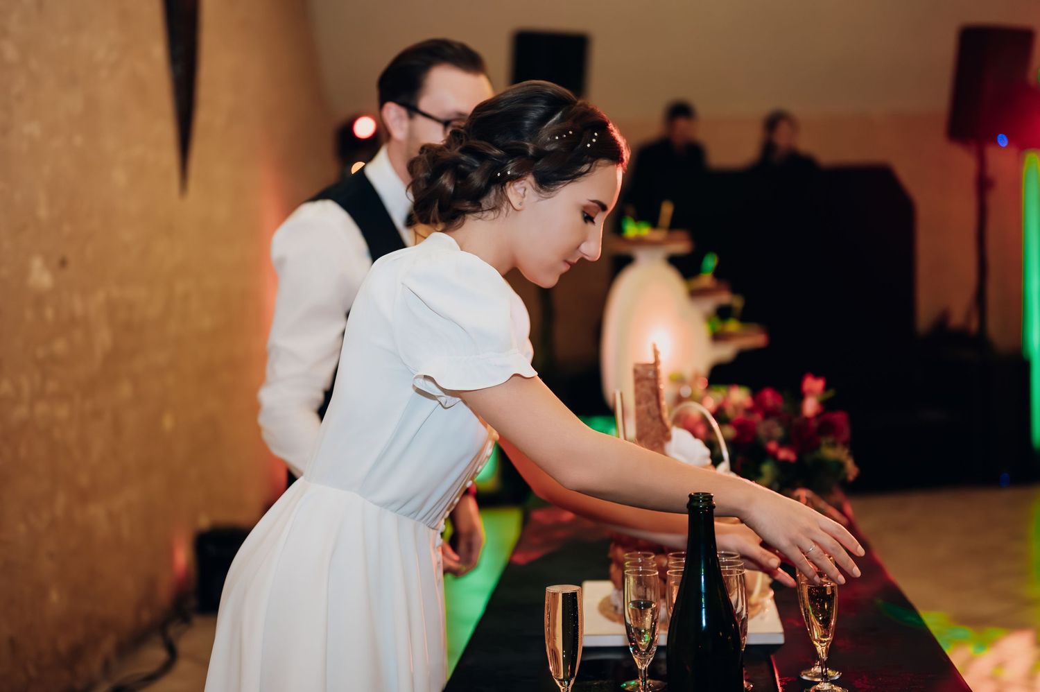 Bride in white dress arranging champagne glasses at wedding reception bar table.