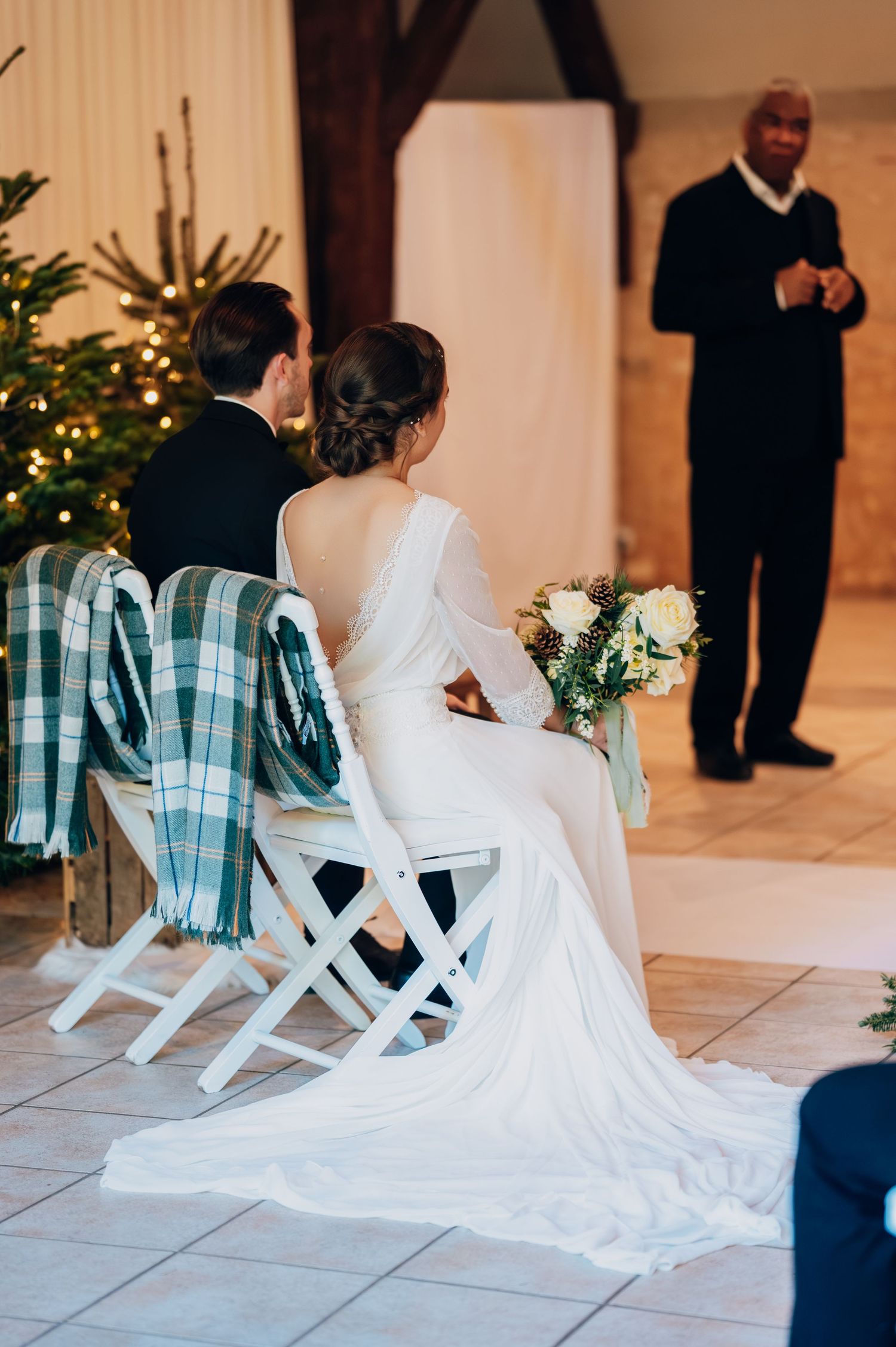 Bride in backless gown and groom seated at altar as officiant addresses them in festive barn venue.
