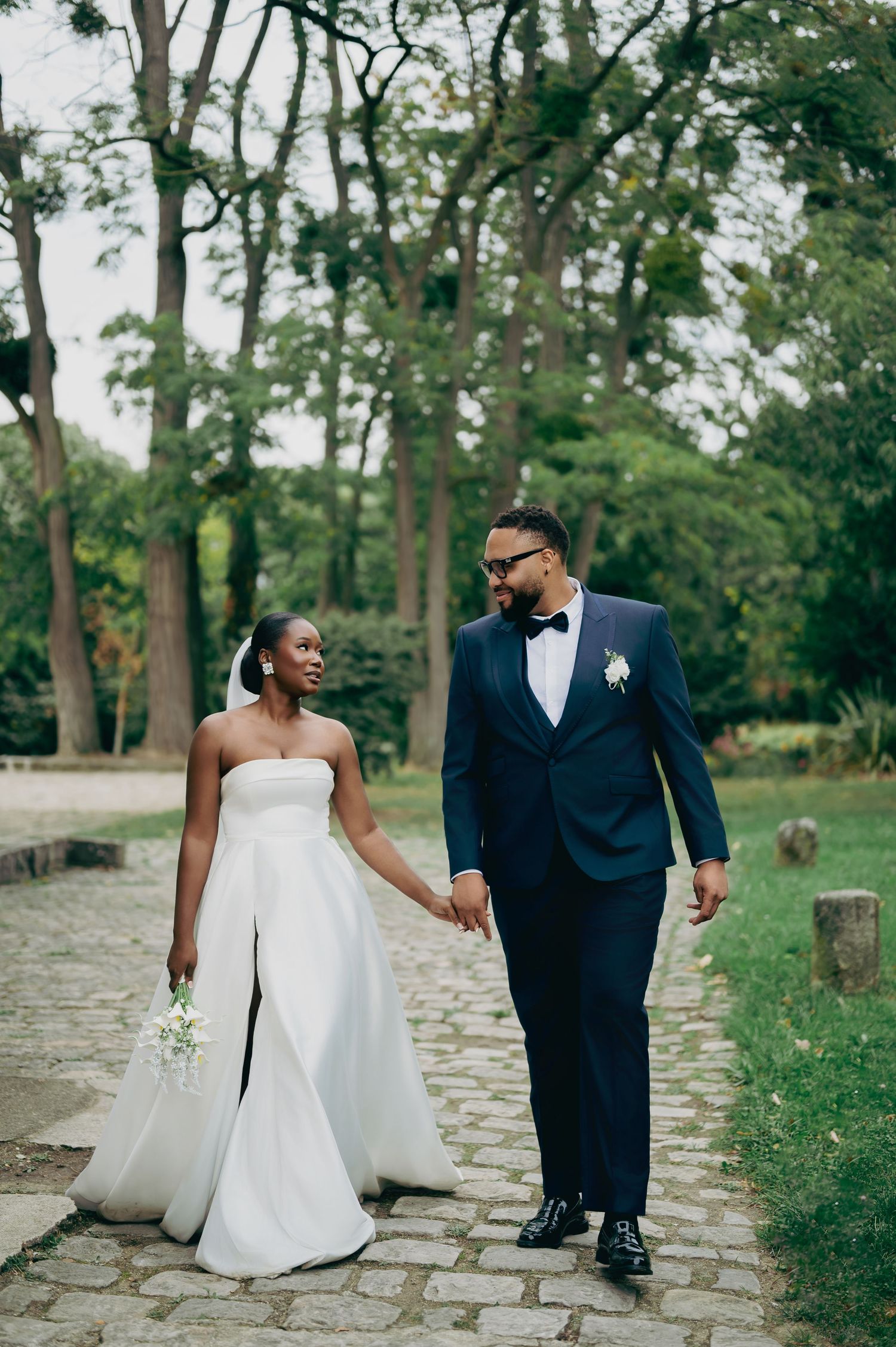 Black bride and groom walking hand-in-hand on cobblestone path, she in white strapless gown, he in navy tuxedo.