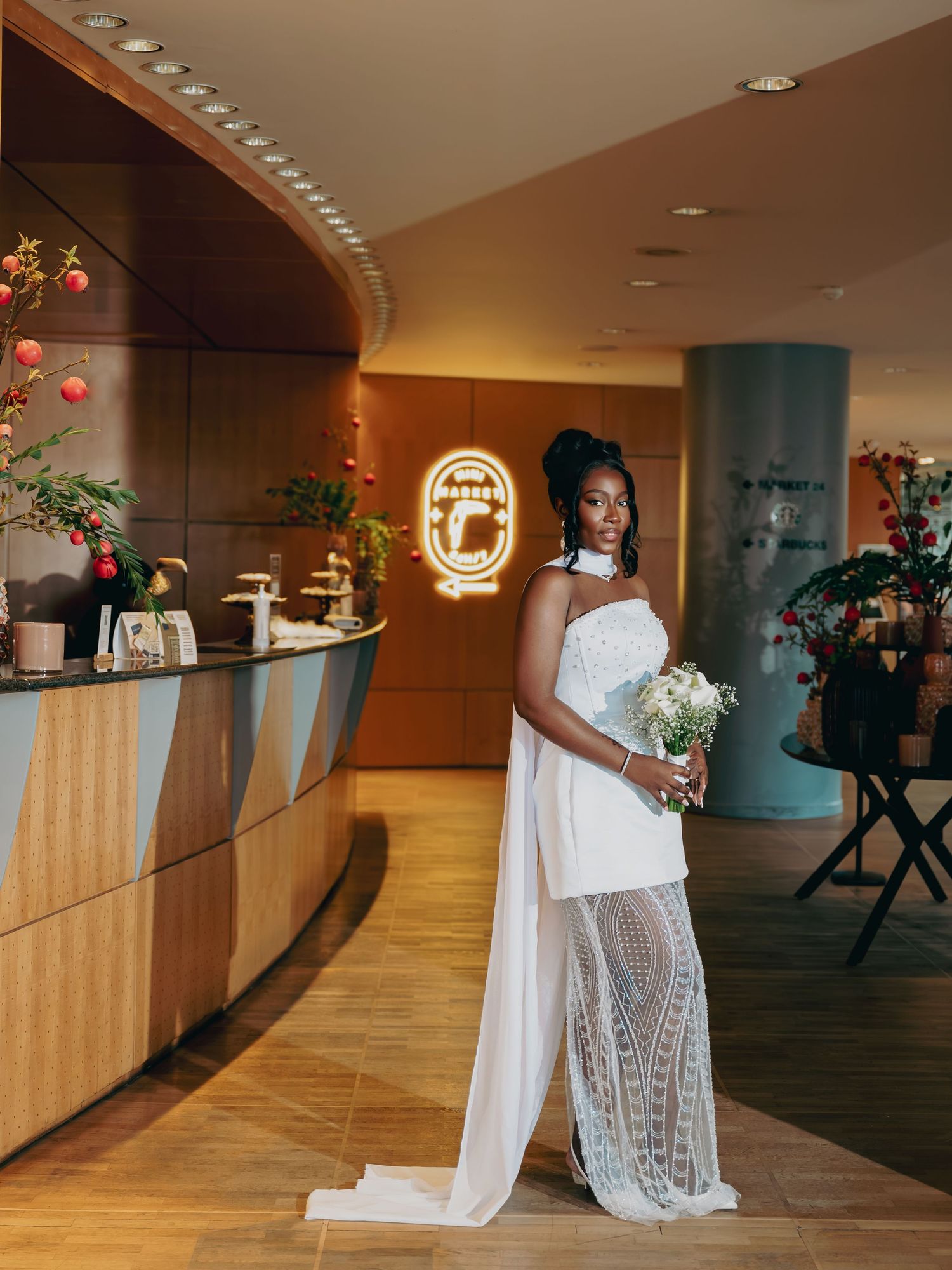 Elegant bride in white lace gown holding bouquet posing in a modern hotel lobby.