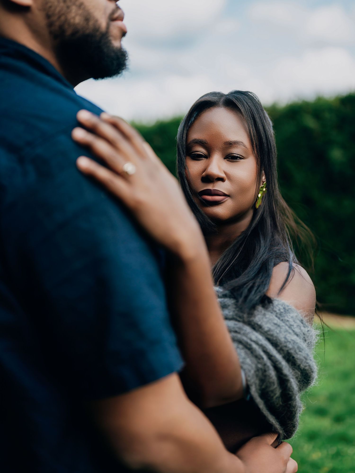 A couple shares an intimate moment outdoors, woman in gray wrap gazes at man wearing engagement ring.