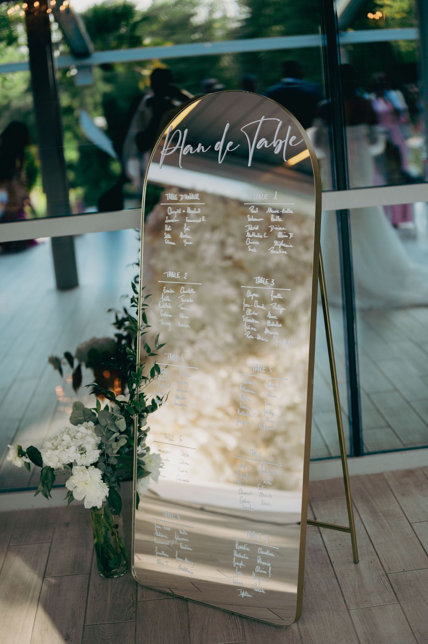 Elegant gold arched mirror seating chart with white calligraphy and floral arrangement at wedding reception.