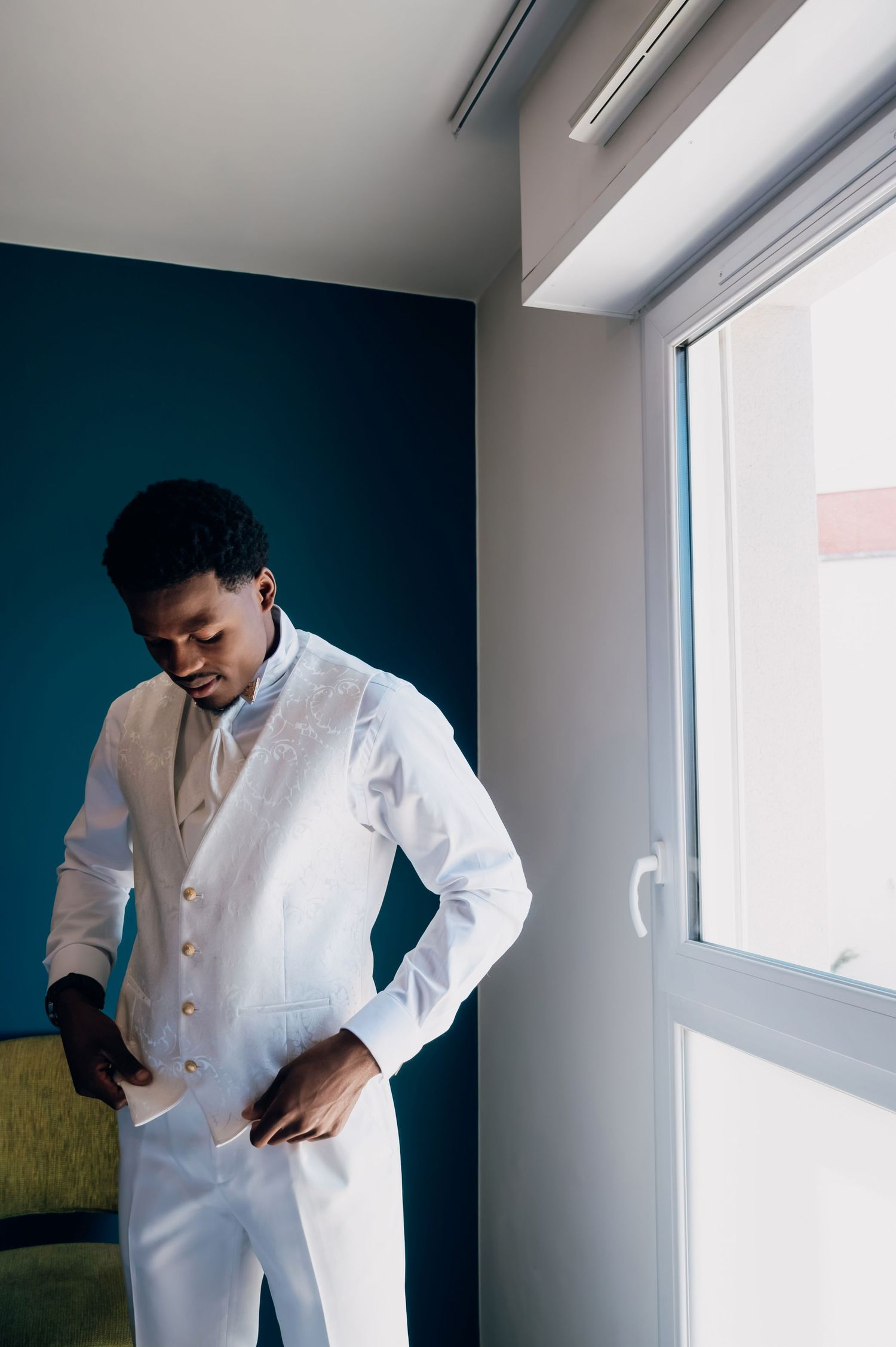 Groom in white suit adjusting his jacket near a window with teal accent wall.