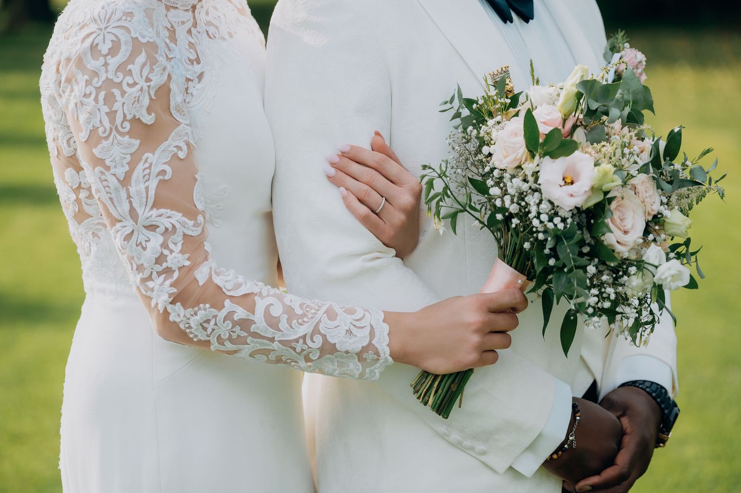 Close-up of interracial couple embracing, bride in lace long-sleeve gown holding a soft pink and white bouquet.
