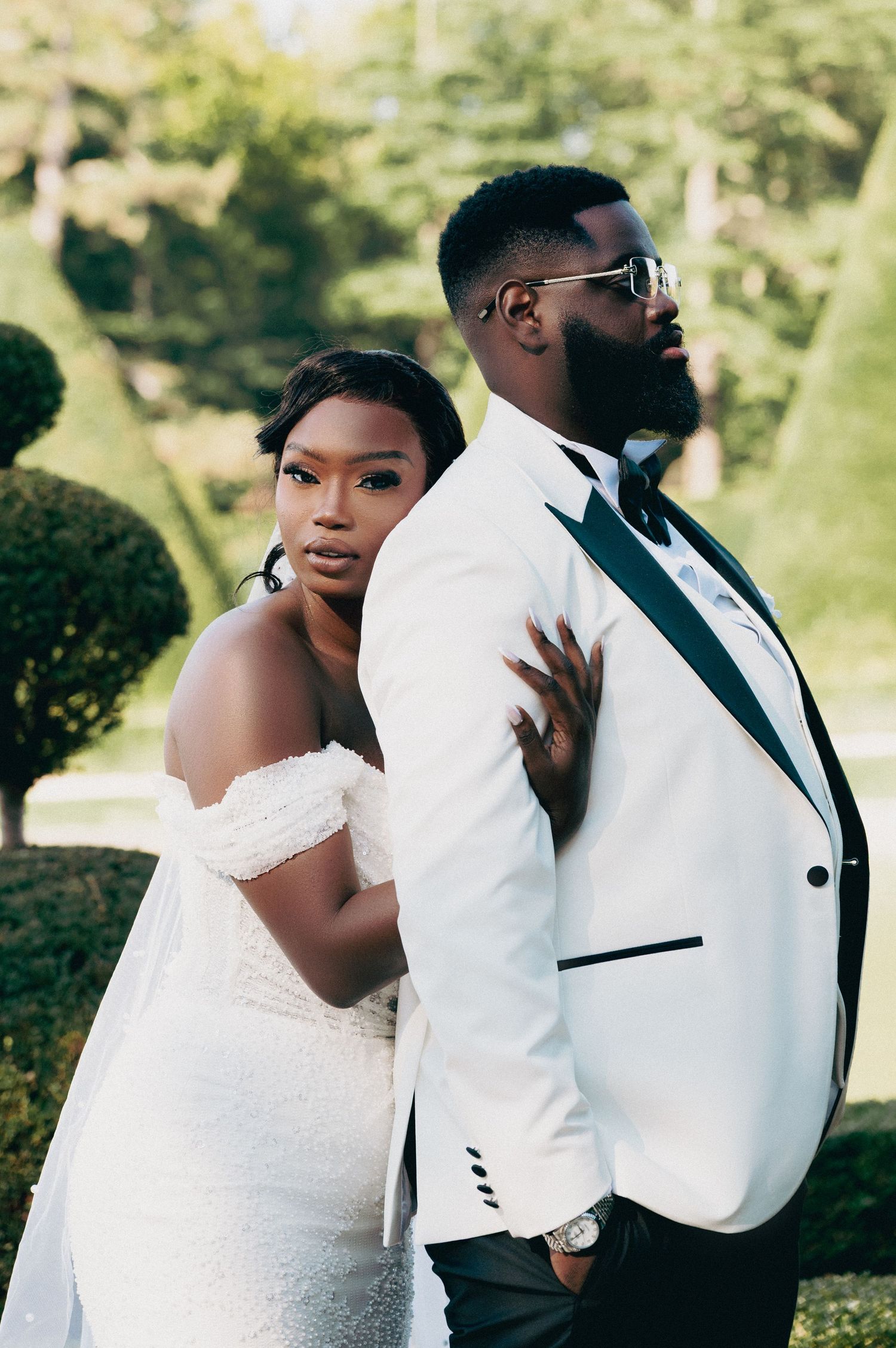 Bride in off-shoulder gown and groom in white tuxedo posing back-to-back in a garden.