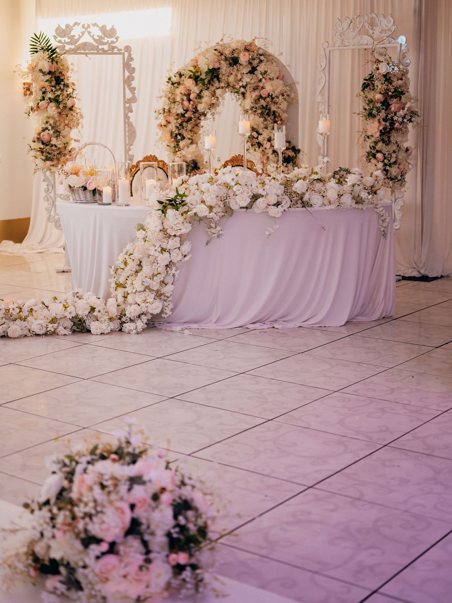 Elegant wedding sweetheart table adorned with lush white and blush pink floral arrangements and a floral arch backdrop.
