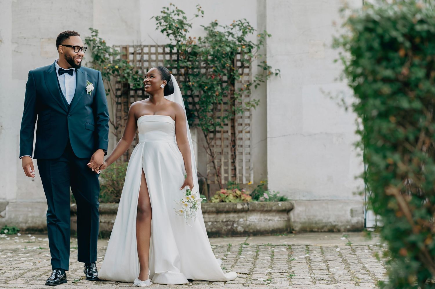 Elegant Black bride and groom in white strapless gown and navy suit holding hands in courtyard.