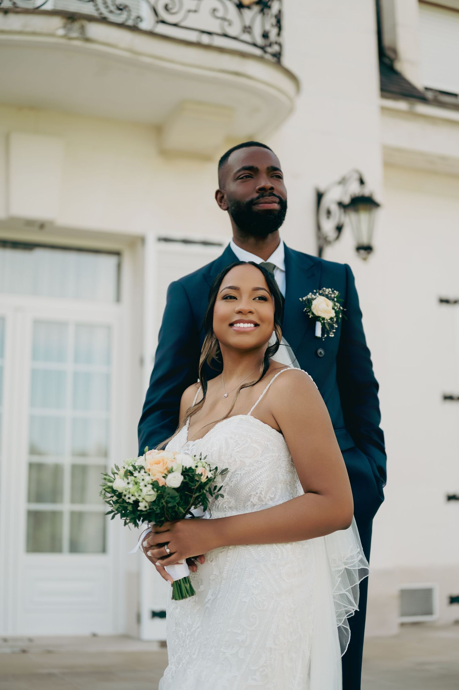Interracial wedding couple pose elegantly outside a white European building, bride holding a white floral bouquet.