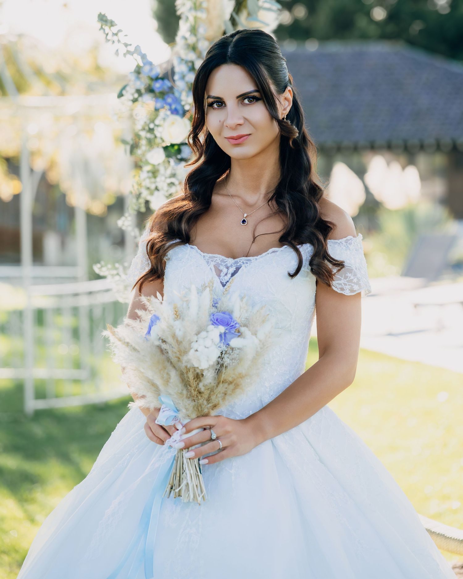 Beautiful bride in off-shoulder white ball gown holding blue and ivory bouquet standing before floral arch outdoors.