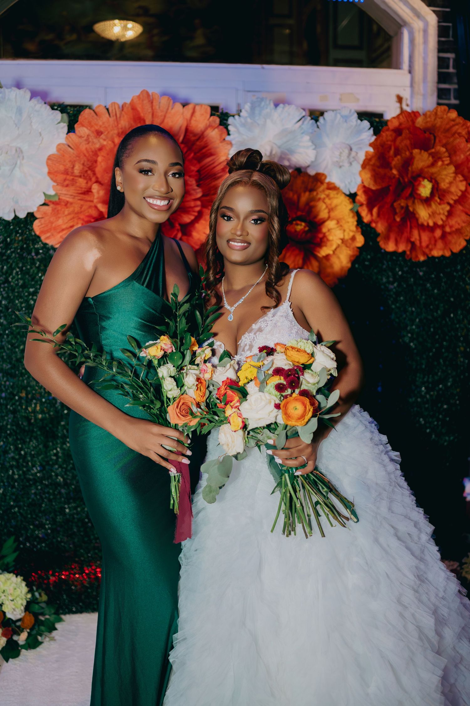 Bride in a white ruffled gown poses with bridesmaid in emerald green dress before a colorful paper flower backdrop.