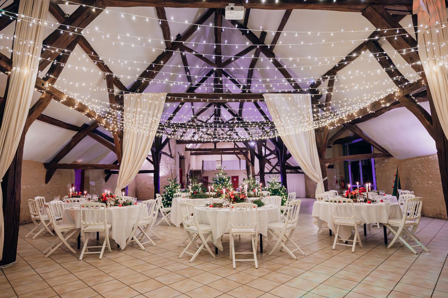Elegant barn wedding reception with fairy lights, white draping, round tables, and white chairs creating a magical atmosphere.