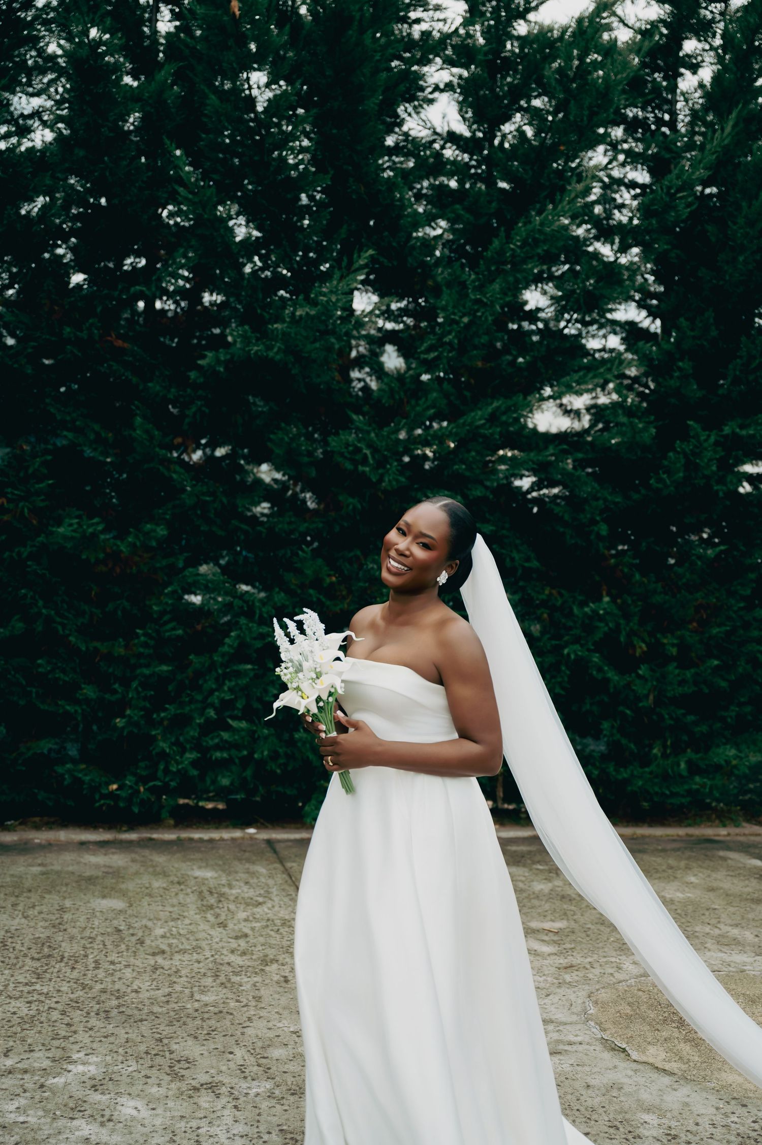 Radiant bride in strapless white gown holding white bouquet with long veil flowing beside tall hedges.