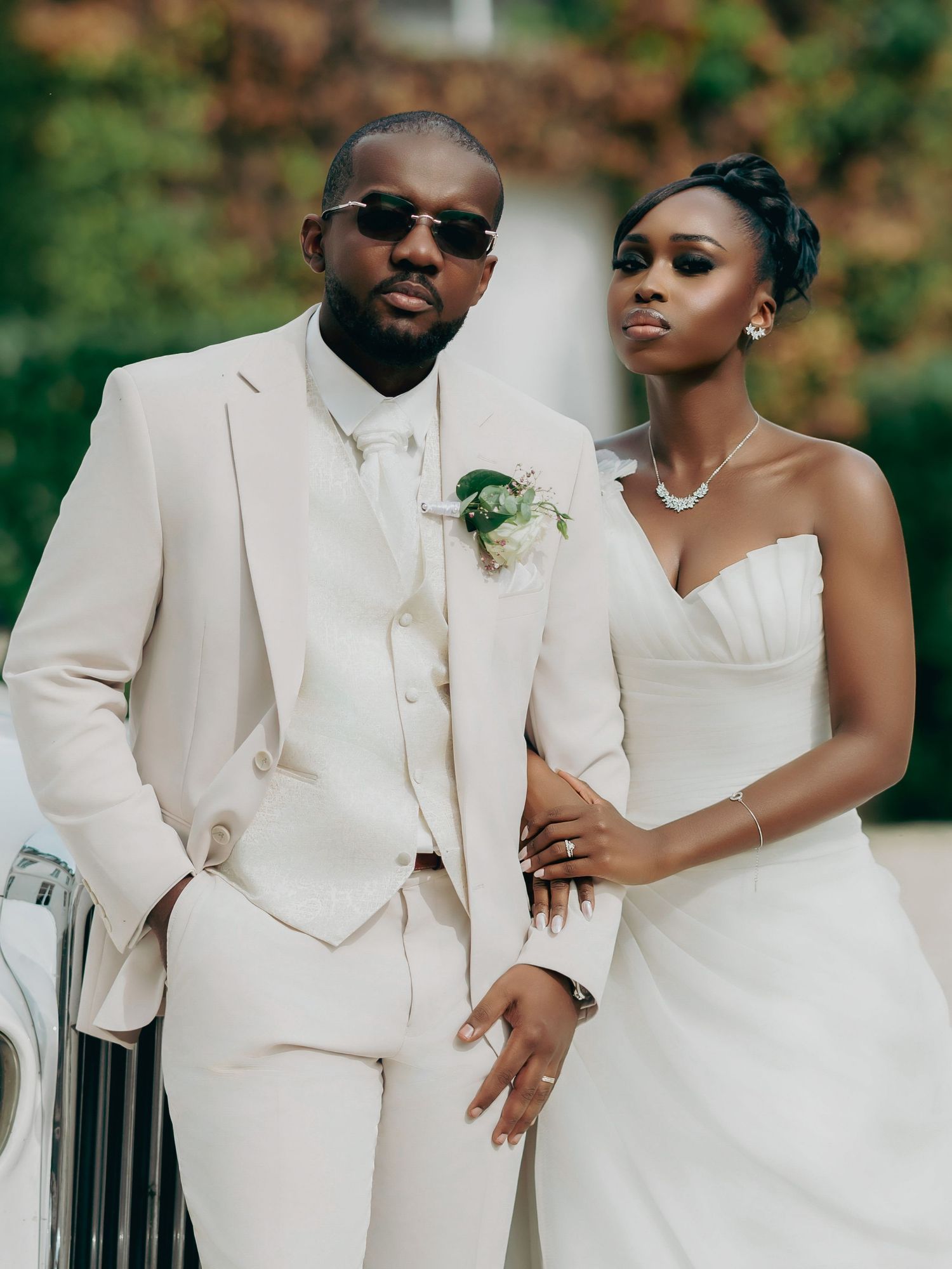 Sophisticated bride and groom in coordinating white wedding outfits posing beside a vintage white car.
