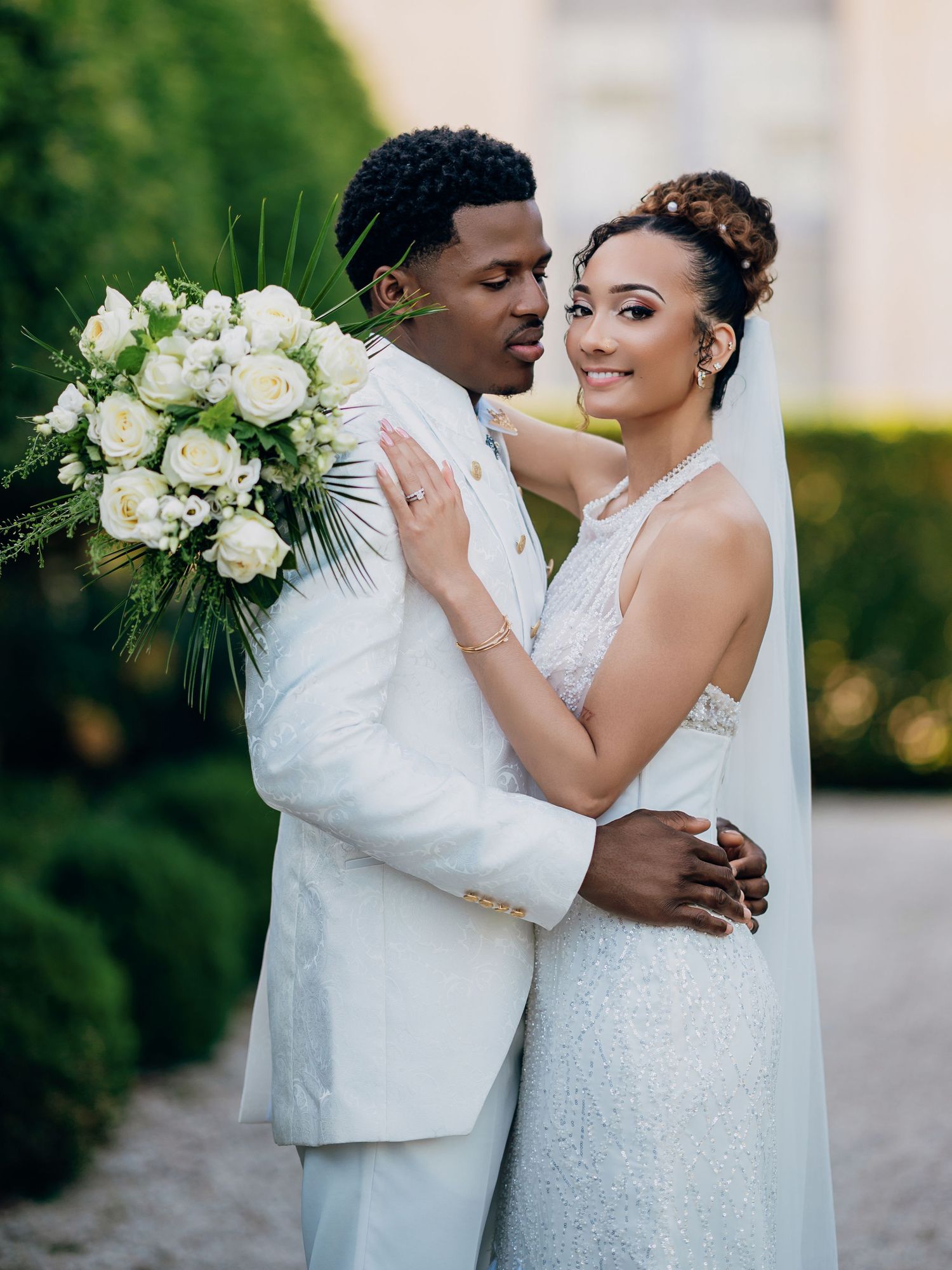 Groom in white military suit embraces bride in lace gown holding white rose bouquet in formal garden.