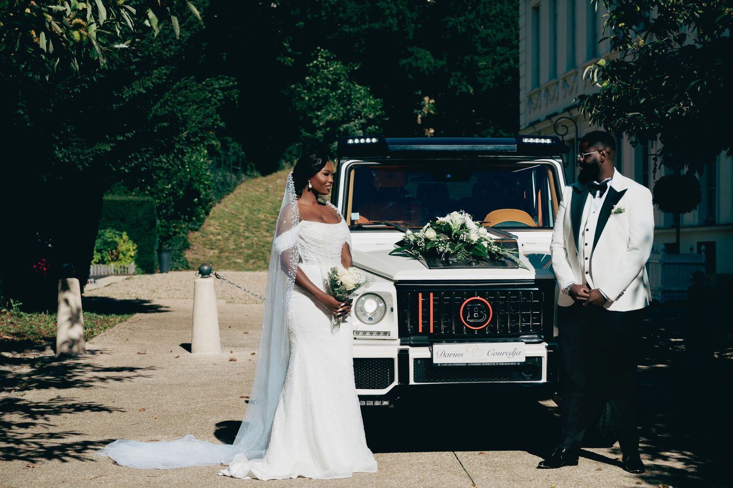 Bride and groom pose beside white Mercedes G-Wagon decorated with flowers on sunny wedding day.