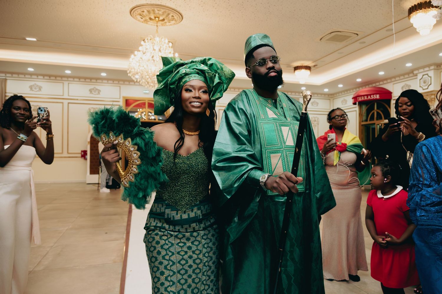 Bride and groom in matching green African traditional outfits with feather fan at wedding reception.