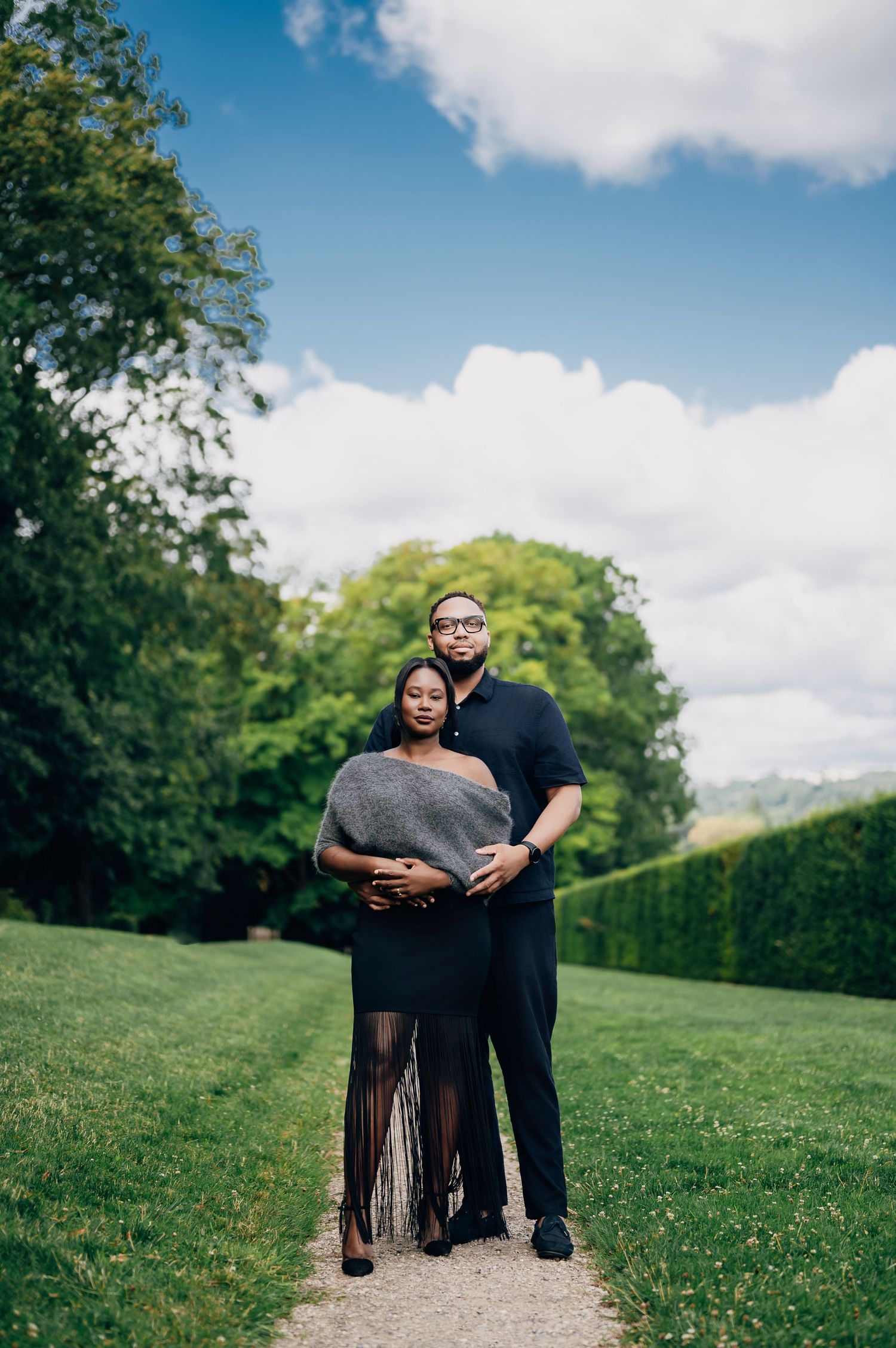 Couple posing together on garden path, man embracing pregnant woman in gray wrap and black fringe skirt.