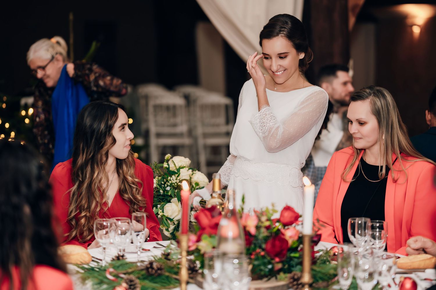 Bride in white dress smiling at wedding reception table with guests in red and pink outfits.