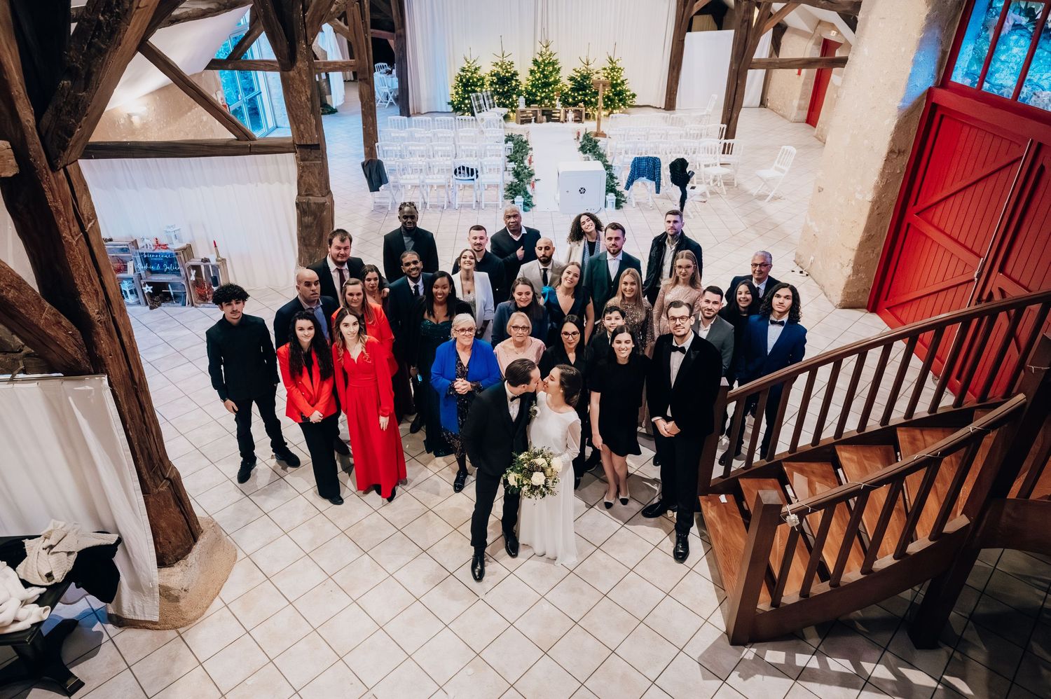 Aerial view of wedding party and guests gathered inside rustic venue with wooden beams and red doors.