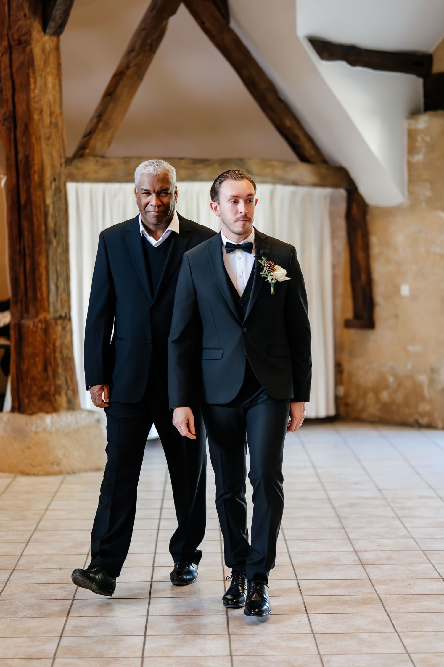 Groom in black tuxedo with bow tie and boutonniere being escorted down the aisle in a rustic barn wedding venue.