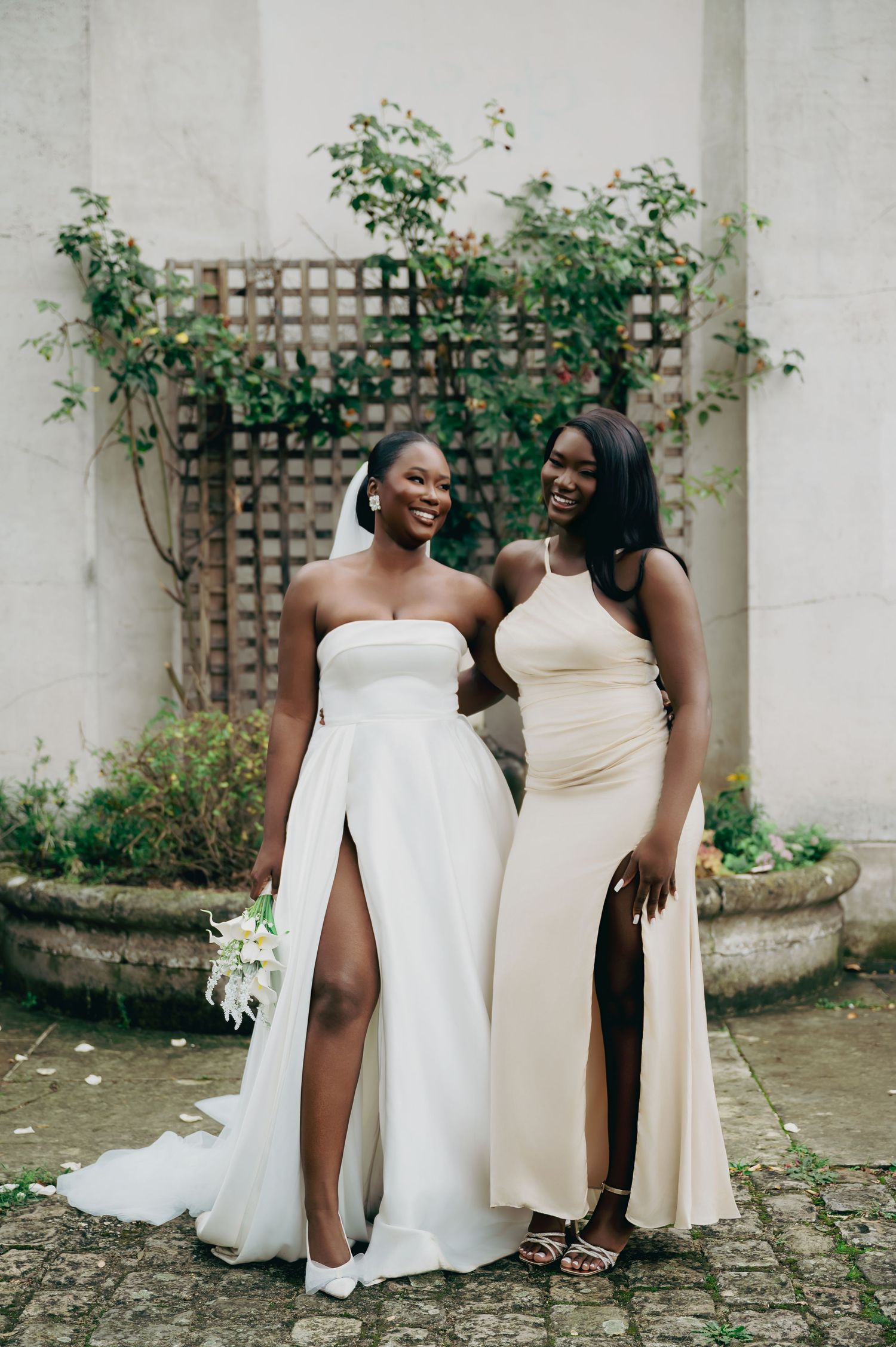 Bride in strapless white gown with slit posing with bridesmaid in champagne halter dress outdoors.