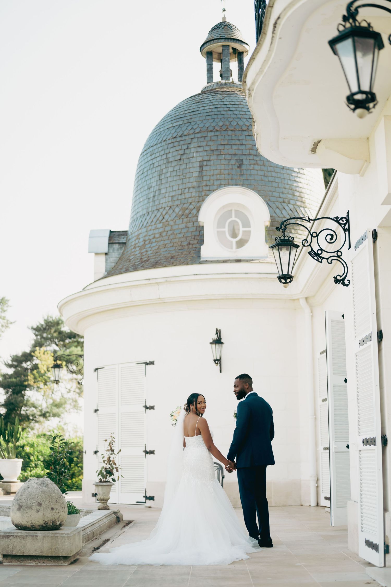Bride and groom hold hands before elegant white château with ornate dome and lanterns.
