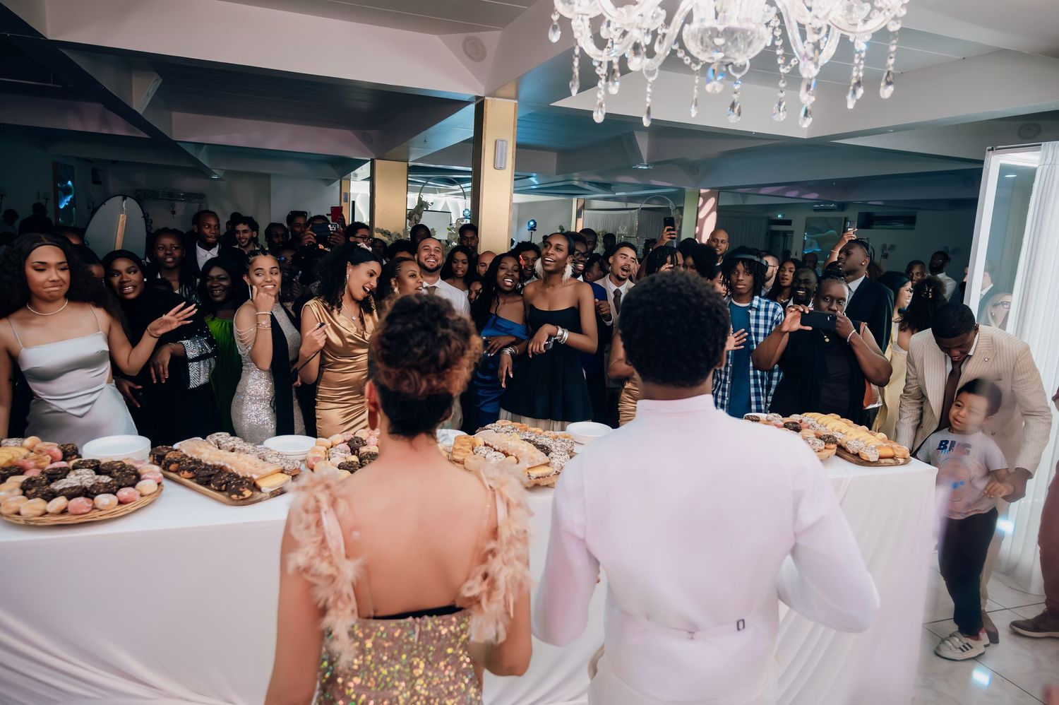 Bride in gold sequin dress and groom in white suit greet guests at crowded wedding reception.