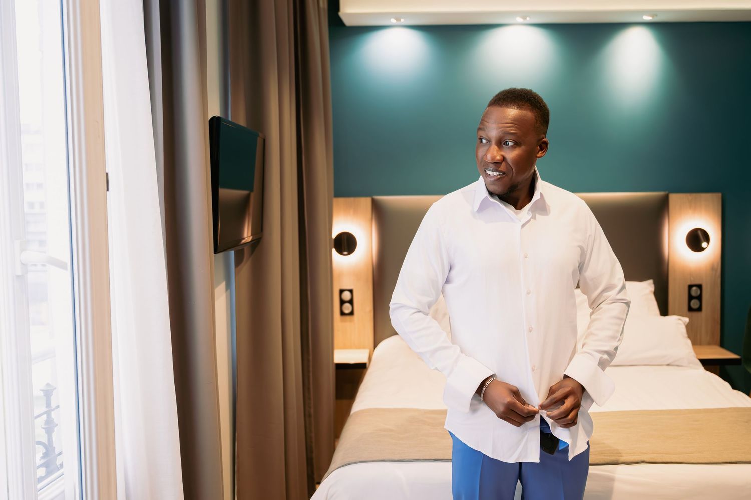 Man in white shirt stands in modern hotel room with teal accent wall.