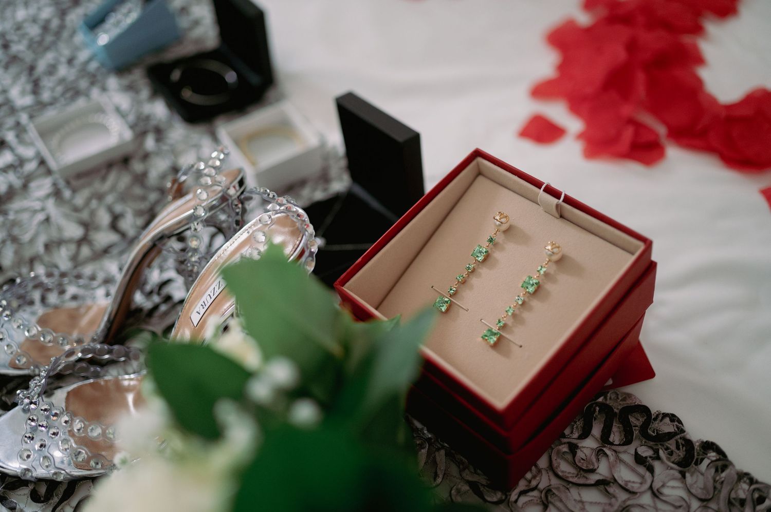 Green gemstone drop earrings displayed in a red jewelry box surrounded by rose petals and bridal accessories.