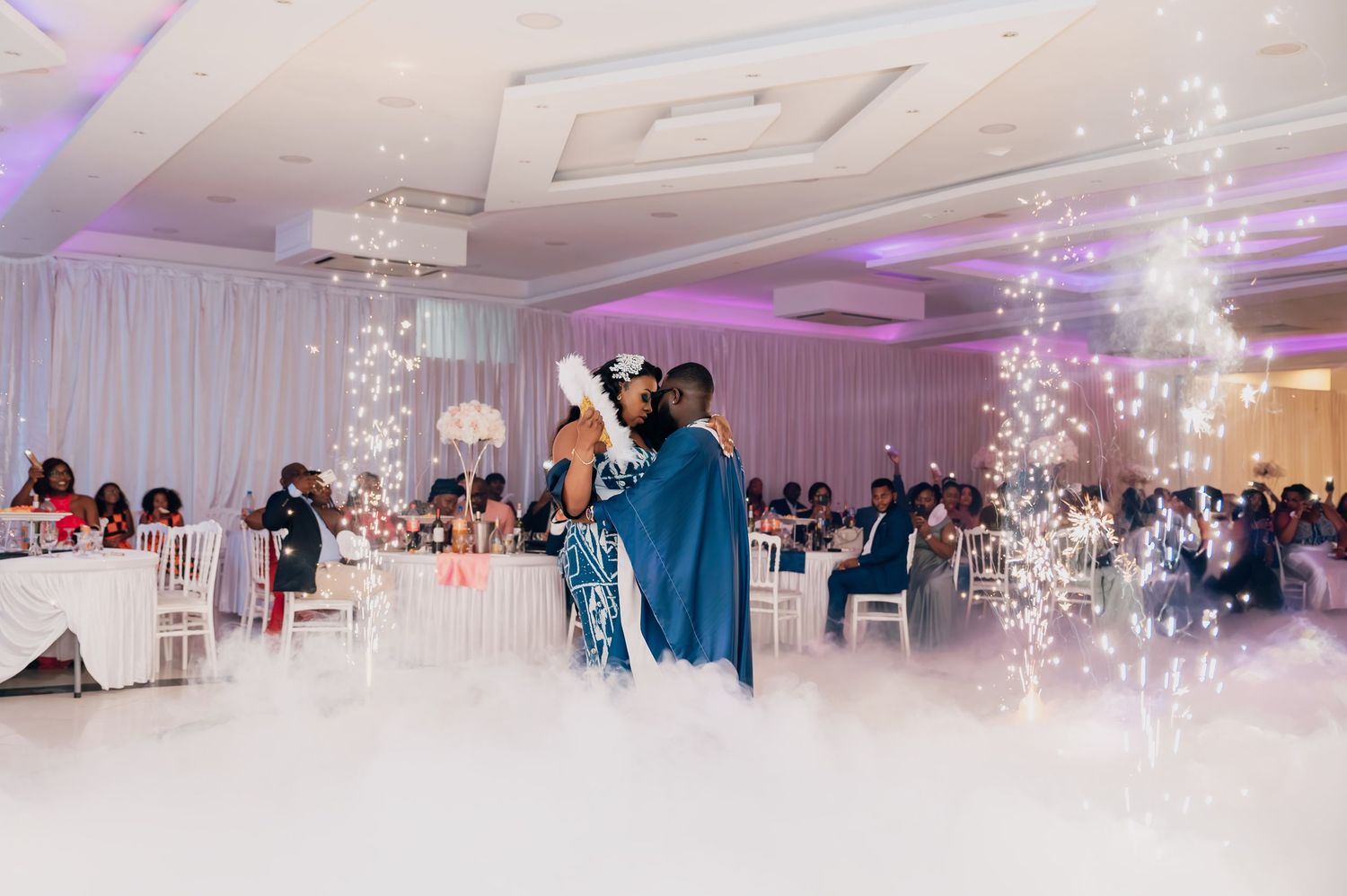 Bride and groom share first dance surrounded by sparkling lights and dry ice fog at reception.