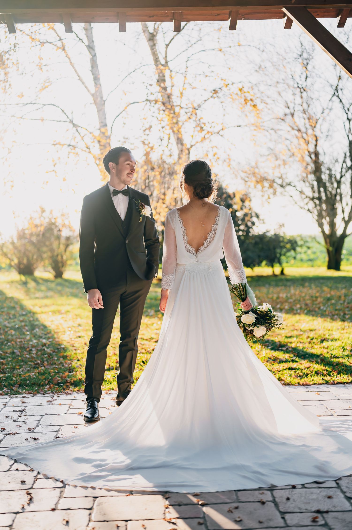 Bride in backless white gown with train walks with smiling groom in black tuxedo through autumn park.