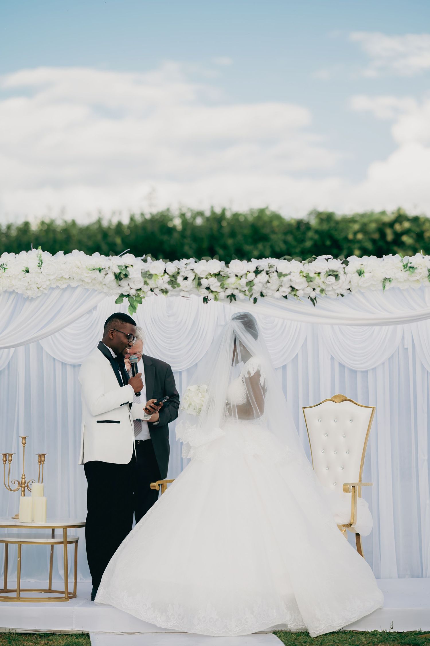 A groom in a white tuxedo reads his vows to his bride before a white floral backdrop at an outdoor wedding.