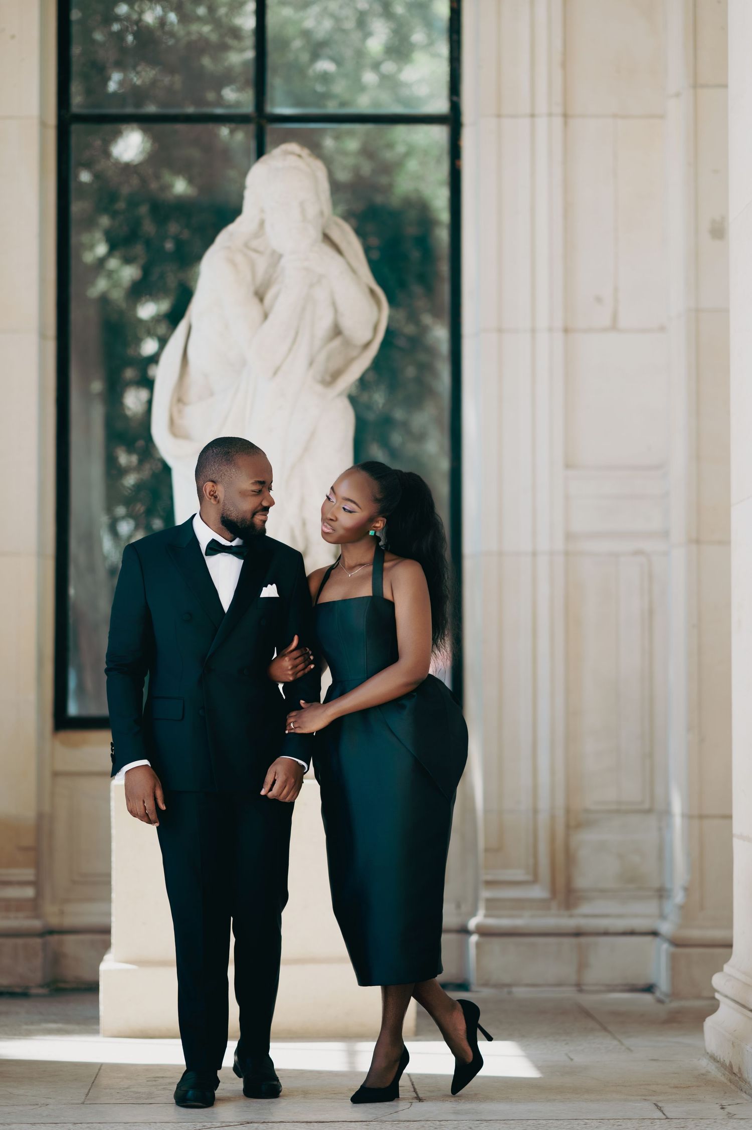 Elegant Black couple in matching dark green formal attire posing in classical building with marble statue backdrop.