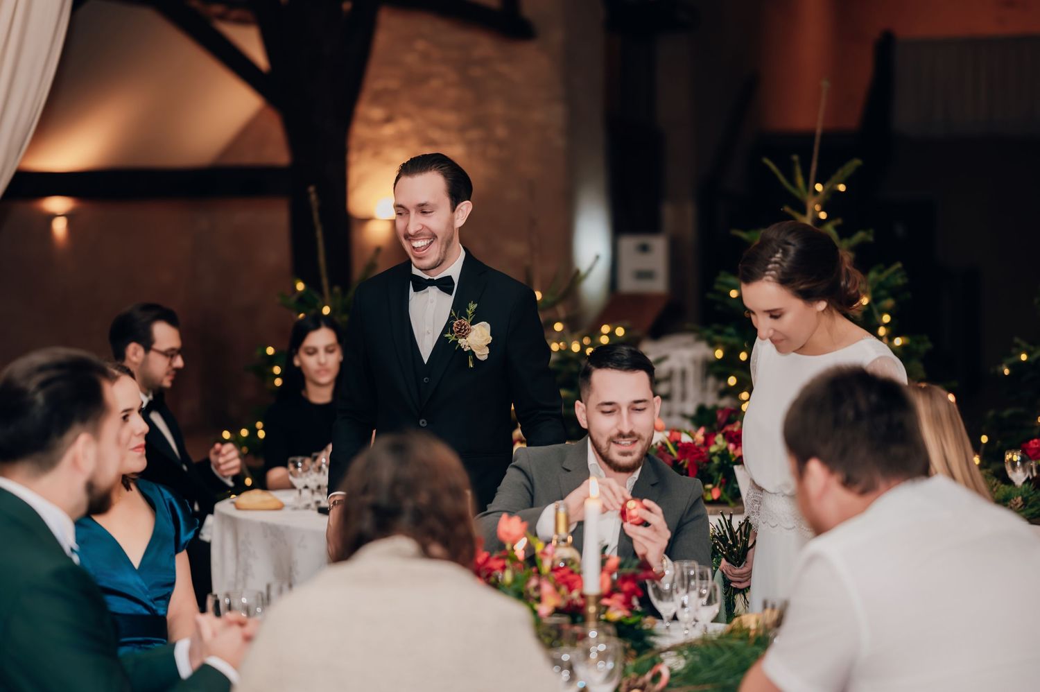 Groom in black suit laughing with wedding guests at festive reception with fairy lights.