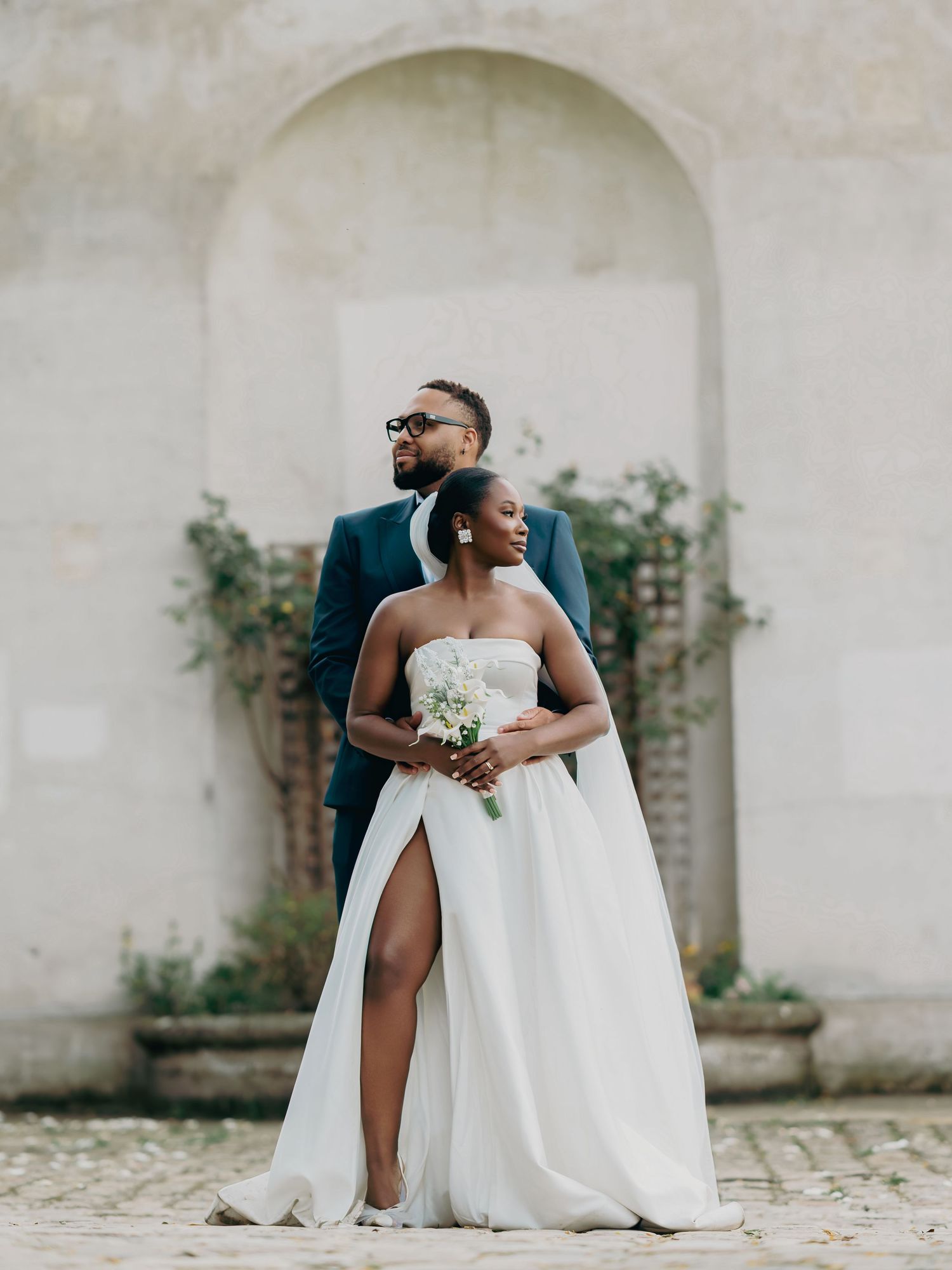 Elegant Black couple posing in wedding attire, bride in strapless white gown with slit, groom in navy suit before stone wall.