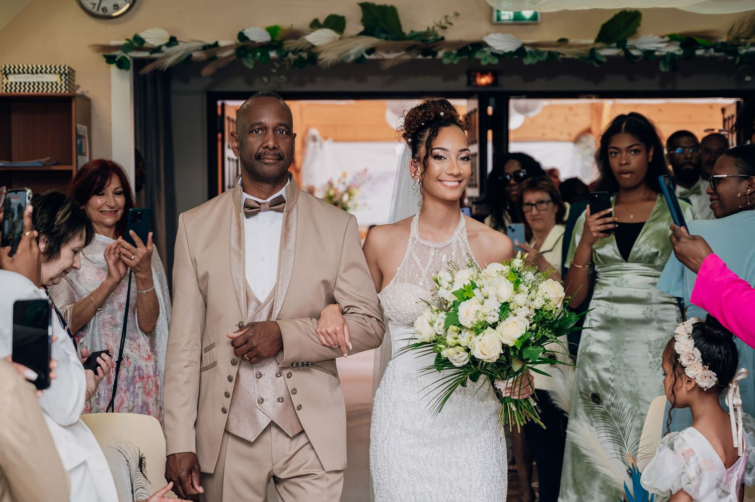 Bride in halter wedding gown walks down aisle with escort holding white bouquet at ceremony.