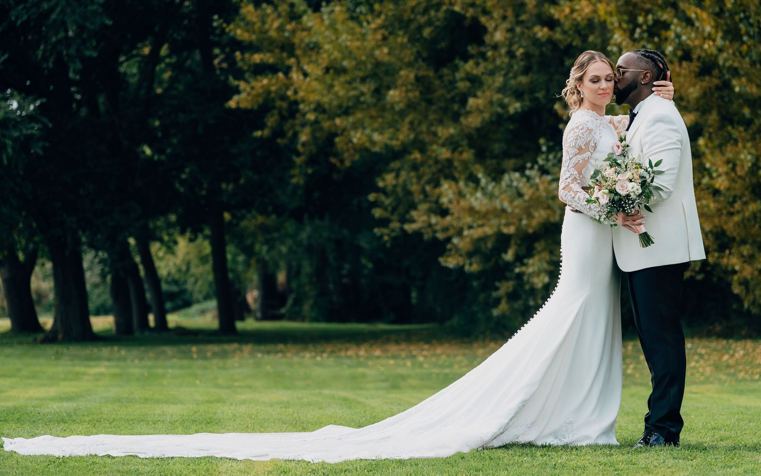 Interracial couple sharing a romantic kiss on a lush green lawn, bride in a long-train white gown with bouquet.