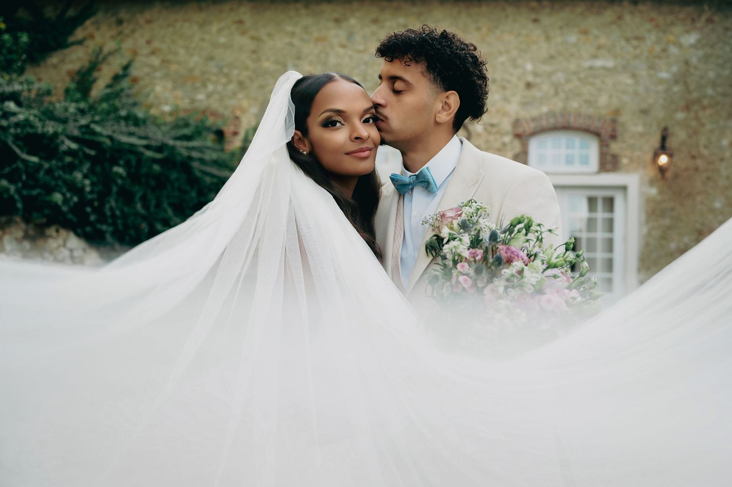 Groom kissing bride on cheek as her long white veil flows in the breeze outdoors.