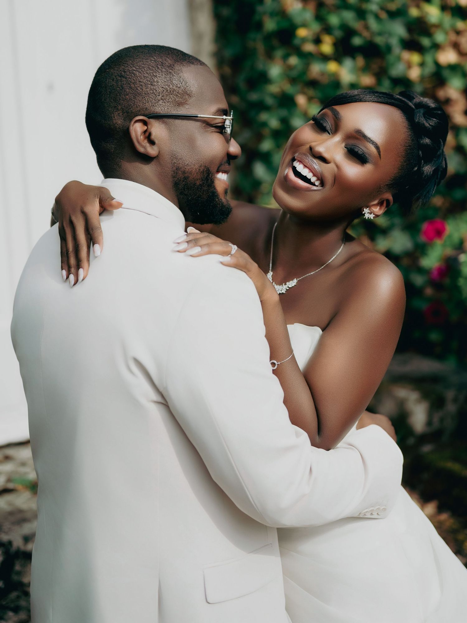 Joyful Black bride and groom embracing outdoors, bride in strapless gown laughing, groom in white suit.