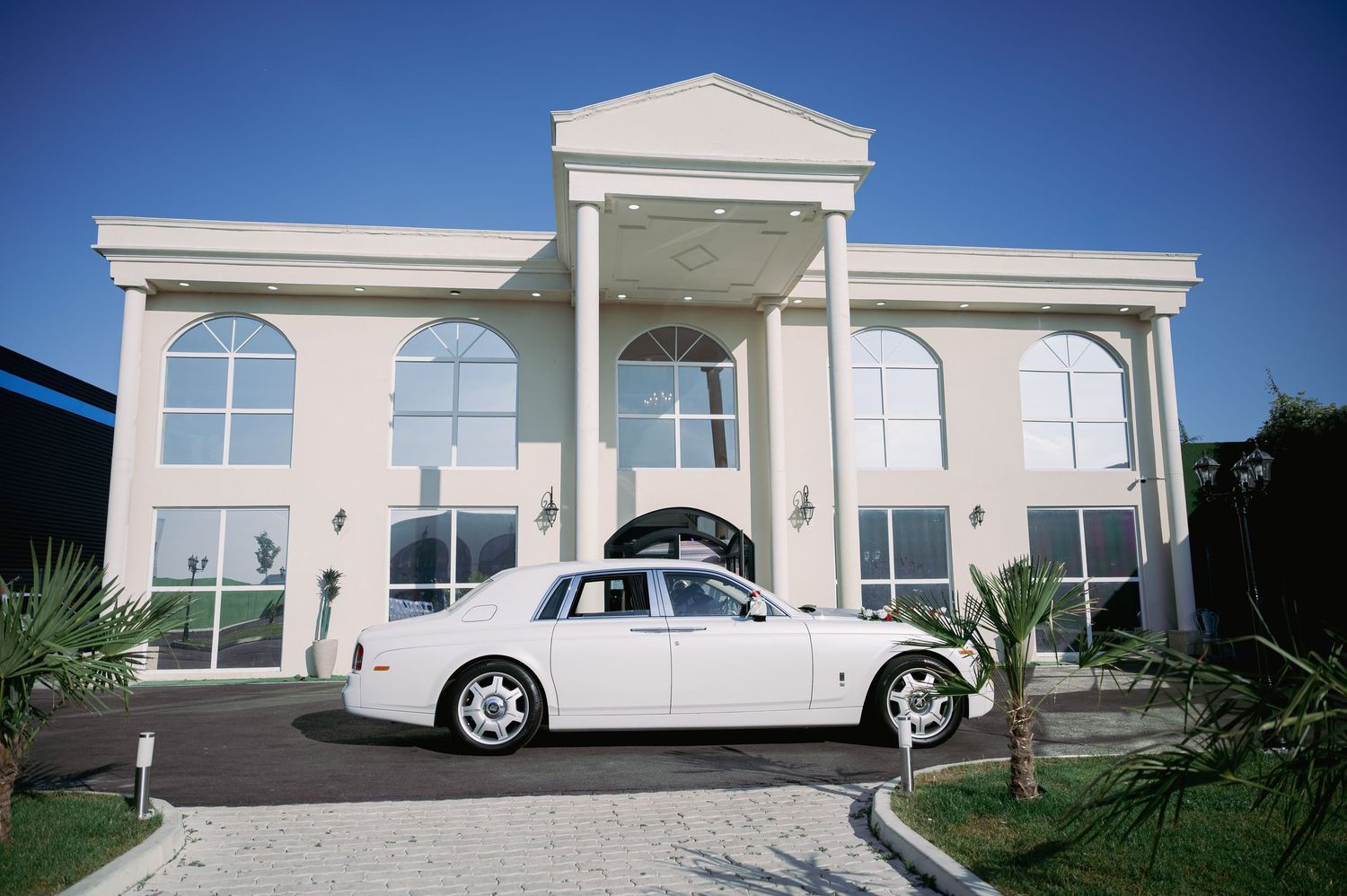 Elegant white Rolls-Royce wedding car parked in front of a luxurious white mansion.