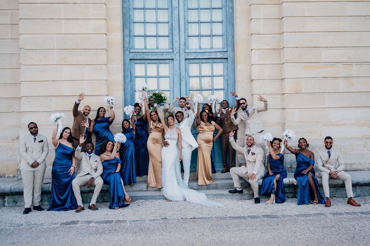 Joyful wedding party celebrating on building steps, bridesmaids in royal blue and groomsmen in champagne suits cheering.