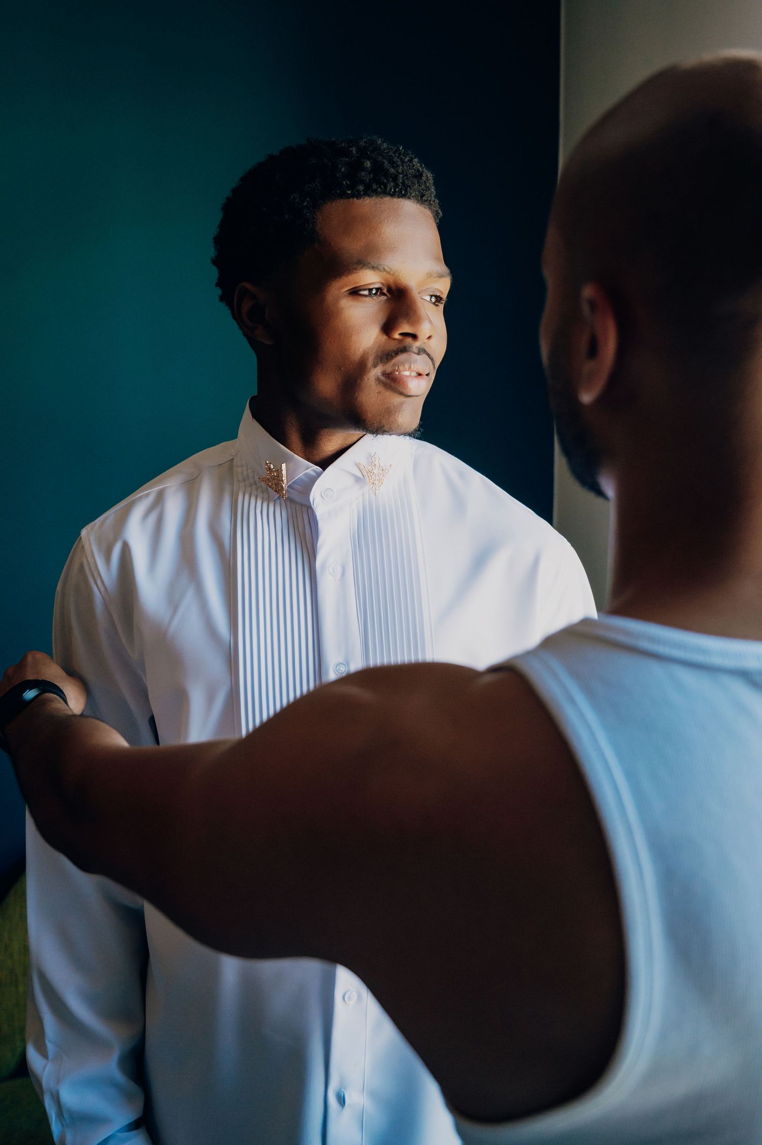Groomsman helping groom adjust white pleated tuxedo shirt before wedding ceremony.