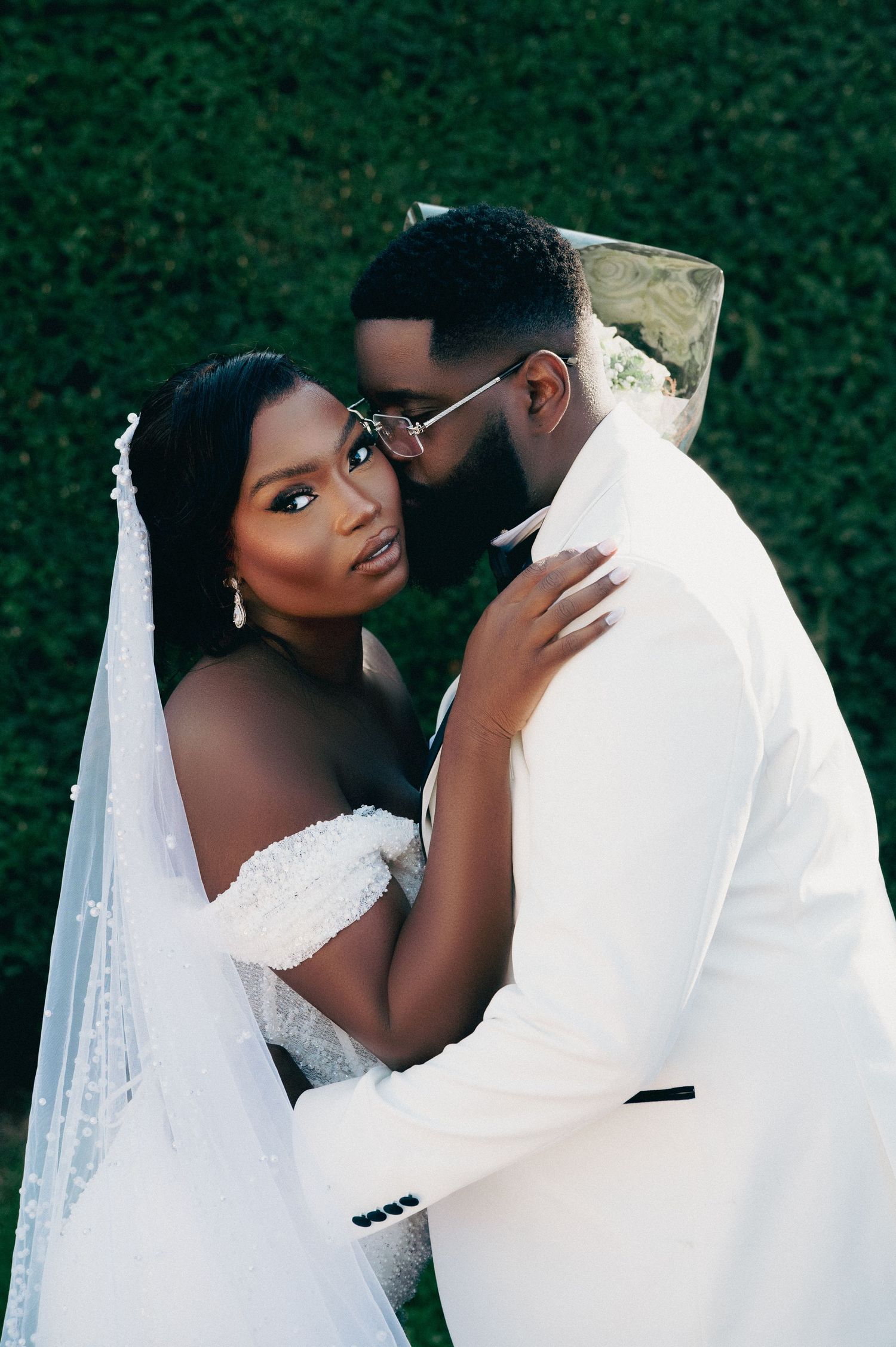 Elegant couple in wedding attire posing closely together against a green hedge backdrop.