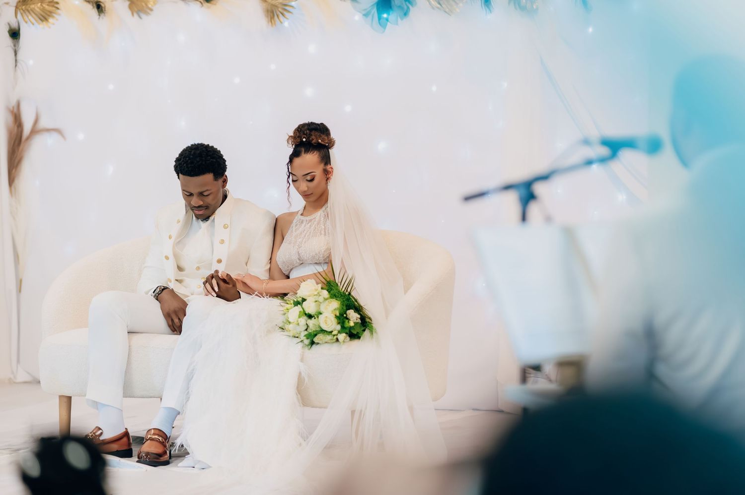 Bride and groom in white outfits sit together on white sofa during wedding ceremony.