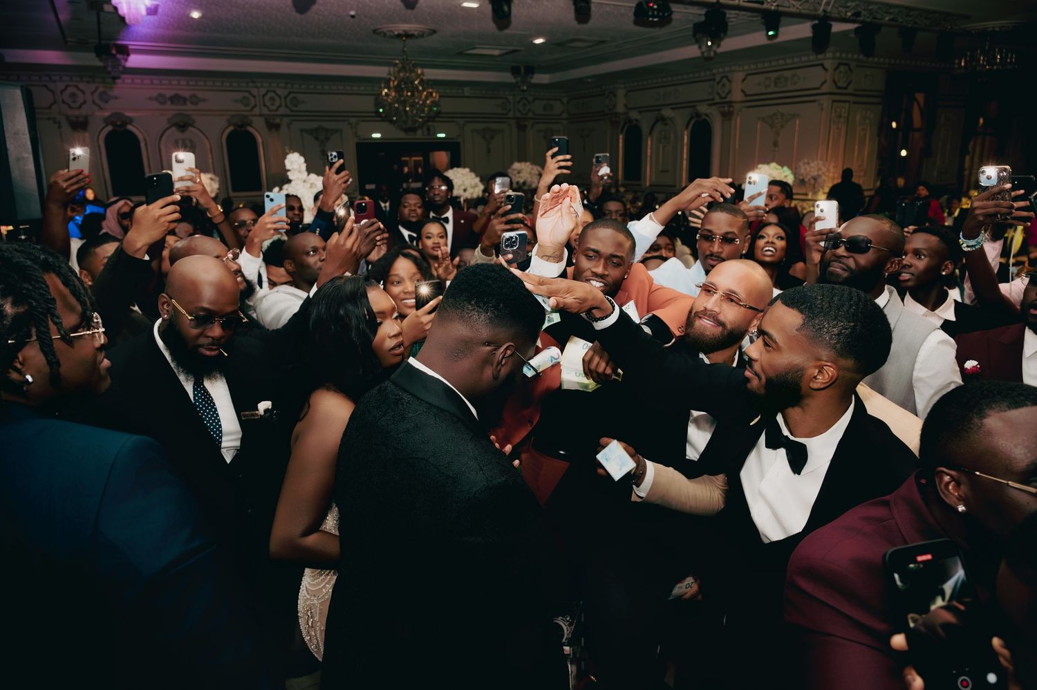 Groom celebrates with groomsmen and guests in lively wedding reception crowd.